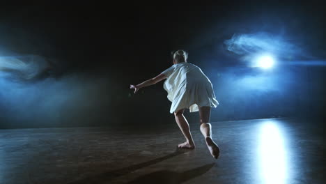 A-young-female-ballerina-barefoot-jumps-on-stage-and-moves-in-slow-motion-in-a-loose-white-dress.